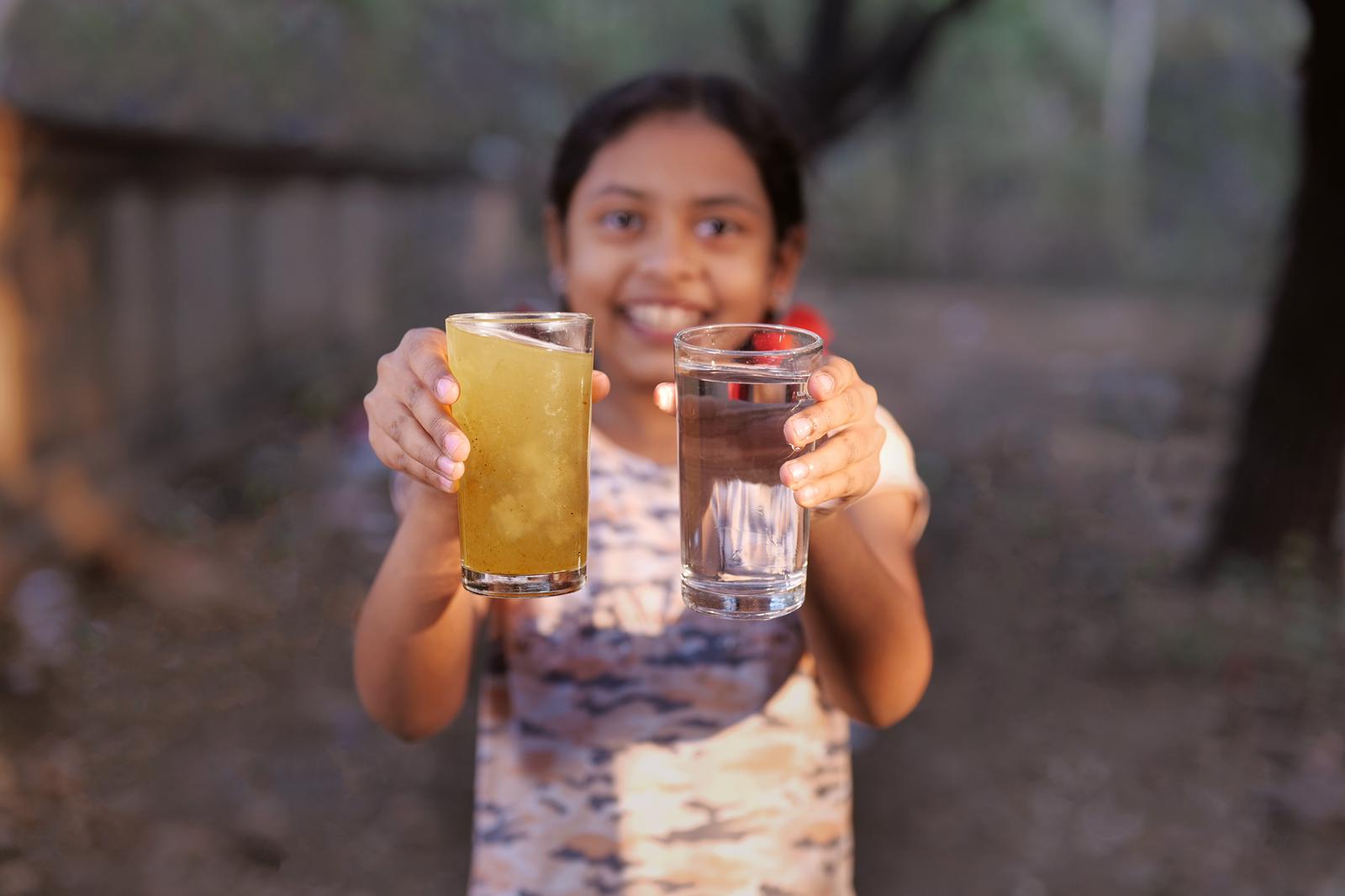 A child in Bela village holds a glass of clean drinking water beside a glass of unsafe water — Nagpur, India.